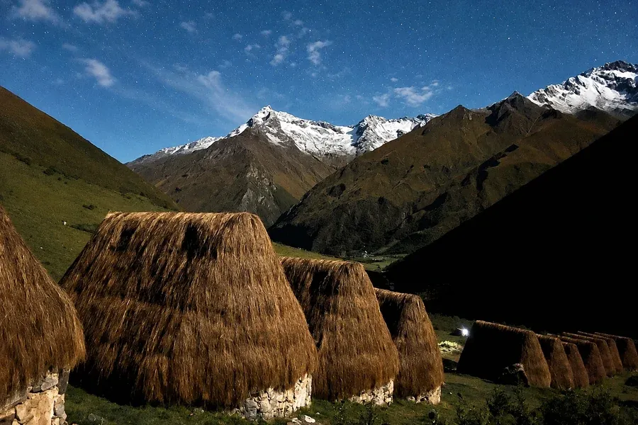 Thatched huts under a starry sky with moonlit snow peaks behind